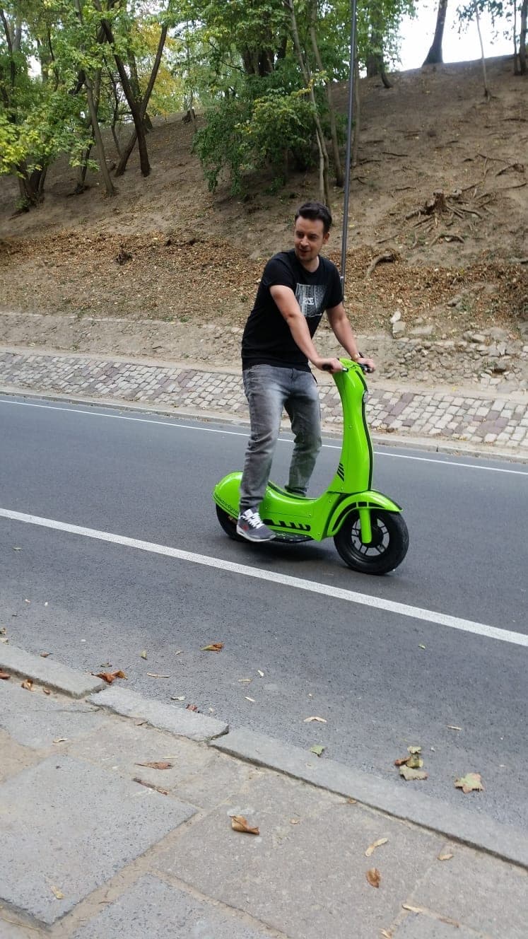 Mobi-2 w jeździe: kierowca stoi obiema stopami na bocznych podestach, w lekkim rozkroku, na drodze wzdłuż parku.