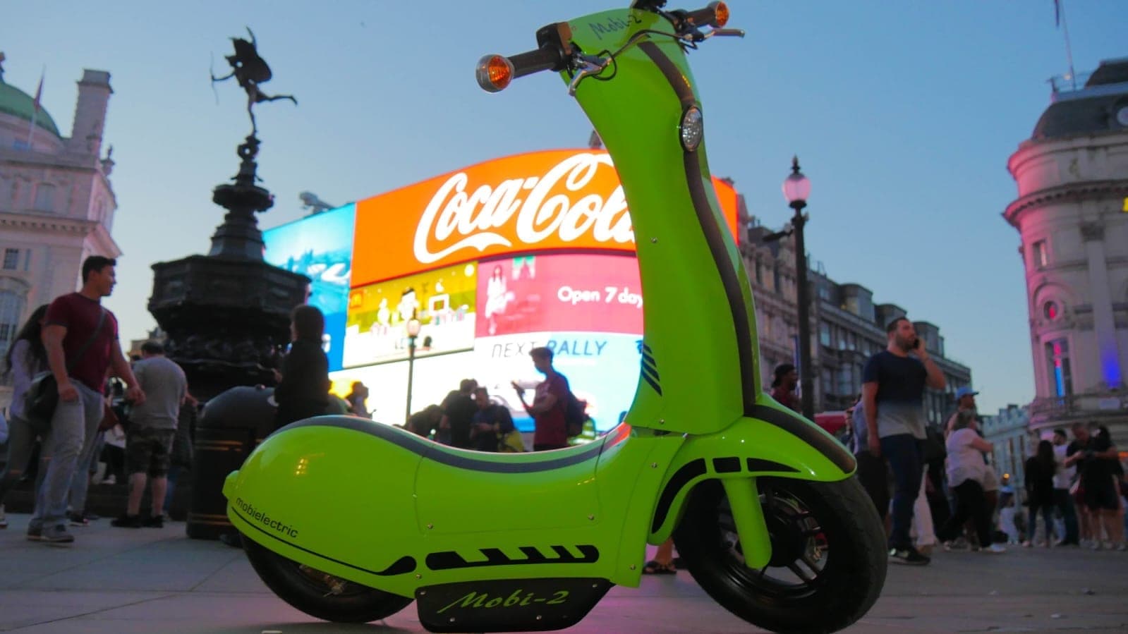 Mobi-2 z boku na Piccadilly Circus o zmierzchu: cała sylwetka z kierownicą i reflektorem, w tle posąg Erosa i rozświetlone billboardy.